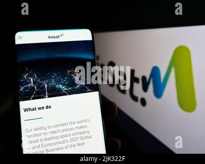 Person holding smartphone with webpage of US communications company Viasat Inc. on screen in front of with logo. Focus on center of phone display. Stock Photo