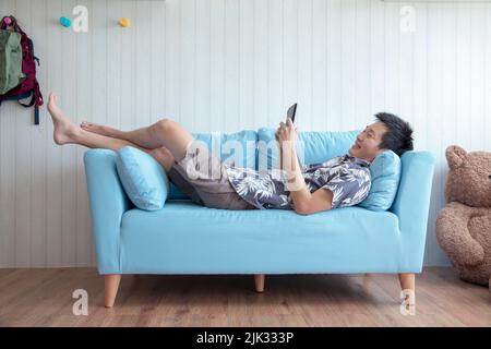 Happy young man lying on couch playing tablet at home in living room Stock Photo