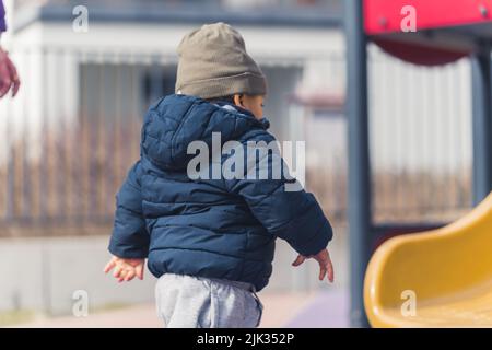 A cute african american male child dressed in a winter clothing walking in a playground area towards the colourful slide - closeup. High quality photo Stock Photo