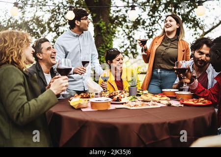 Group of multiethnic carefree friends talk, joke and drink wine during the brunch of the harvest wine tasting, sitting at the table on the terrace of Stock Photo