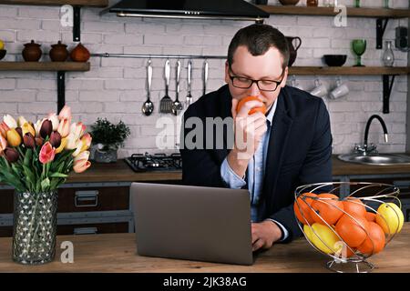 Portrait of a young successful businessman using a laptop sitting at home in the kitchen in a business suit wearing glasses. Man enjoys the smell of a Stock Photo