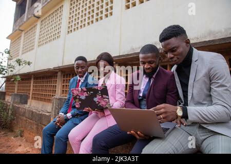 Four young adults sitting on the small wall discuss working topics using  their laptops Stock Photo