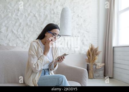 A young beautiful woman at home is sitting on the sofa in the living room, smiling joyfully, holding a smartphone in her hands, receiving the joyful news of confirmation of admission to university Stock Photo