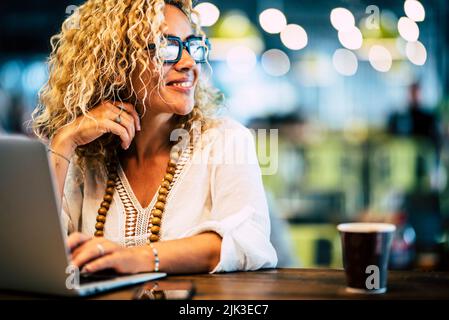 Attractive woman smile and look around while work on laptop sitting at the bar. Modern female people working online. Digital traveler nomad lifestyle. Stock Photo