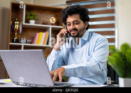 Happy businessman talking with client on mobile phone while working laptop at office - concept of consultation, financial advisor and white collar Stock Photo