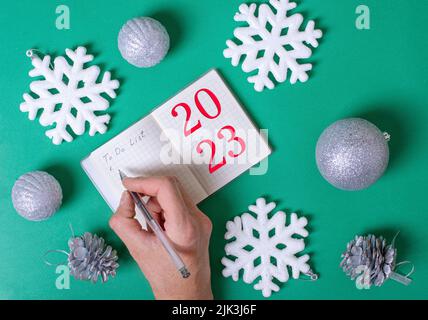 Female hand writes a to-do list in a diary with the inscription 2023 to be done in the New Year 2023 on a green background with silver balls, white sn Stock Photo