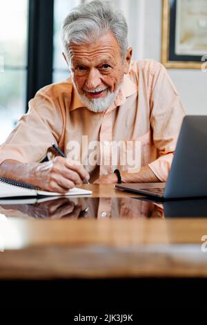 A happy senior student is sitting at home and writing down notes in a notebook while following an online lecture on the laptop. Stock Photo