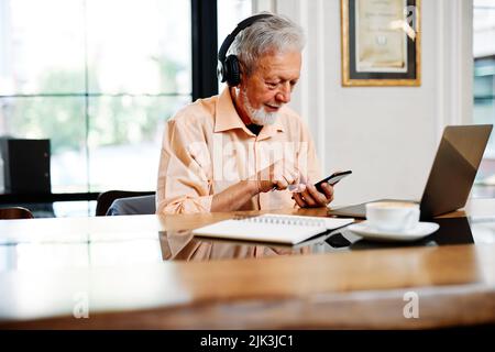 A dedicated senior student is sitting in a cafeteria with headphones on and listening online lecture on the laptop. Stock Photo