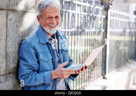 A happy senior student is standing against the brick wall and holding a laptop. Stock Photo