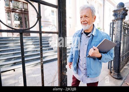 A happy senior student on the retraining program stands in front of the university with a laptop and coffee in his hands. It is never too late to have Stock Photo