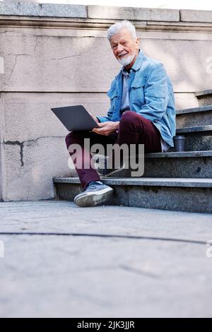 A happy senior student is sitting on the stairs in front of the university building with a laptop in his hands and studying. Retraining concept. Stock Photo