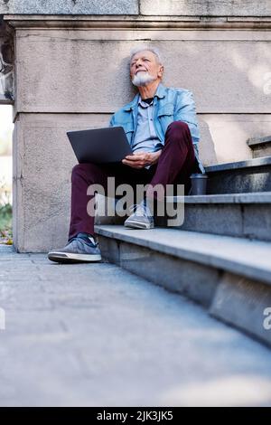 A senior student sits on the stairs of the university building and uses his laptop to look at notes before an exam. Stock Photo