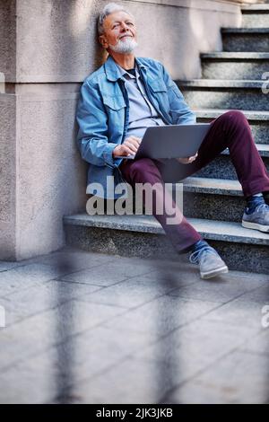 A senior student sits on the stairs of the university building and uses his laptop to look at notes before an exam. Stock Photo