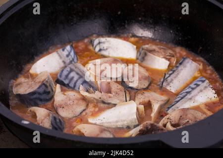 Pieces of mackerel with vegetables are cooked in a cast-iron cauldron. Cooking outdoors. Close-up. Stock Photo