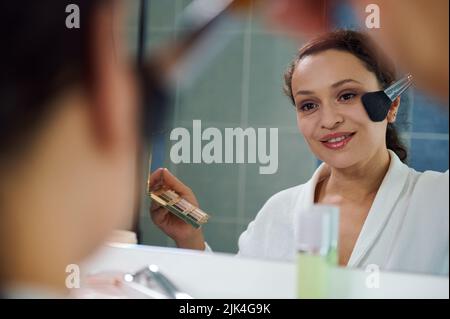 Delightful stunning young woman applies a mattifying powder on her face, satisfied of her appearance and natural make-up Stock Photo