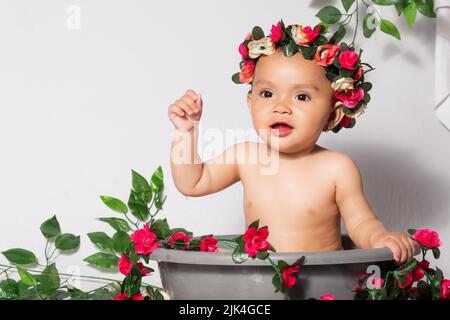 beautiful latina baby girl with brown skin, very smiling and happy with a crown of flowers in a bucket surrounded by flowers. girl sitting holding up Stock Photo