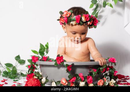 beautiful baby latina with brown skin, inside a gray punch bowl or bucket, surrounded by flowers and roses and red, with a white background. looking d Stock Photo