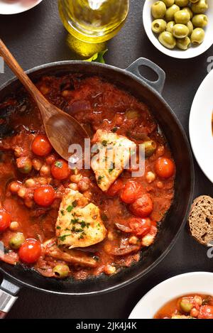 Cod stew with chickpeas, cherry tomatoes and olives in pan over dark stone background. Top view, flat lay Stock Photo