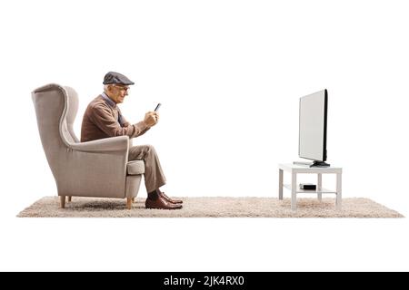 Elderly man in an armchair sitting in front of a TV and using a smartphone isolated on white background Stock Photo