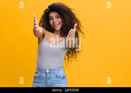 NICE TO MEET YOU. FRIENDLY HUGS CONCEPT. Enjoyed pretty Latin female with afro reach for camera, smile to you, stay isolated over yellow background Stock Photo