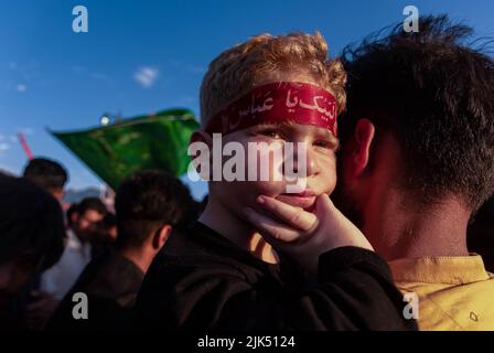 A Kashmiri Shia Muslim boy wearing a headband with Islamic inscriptions looks on towards Shia Muslim men hoisting religious flags and banners as Muharram starts. Muharram is the first month of Islam. It is one of the holiest months on the Islamic calendar. Shia Muslims commemorate Muharram as a month of mourning in remembrance of the Martyrdom of the Islamic Prophet Muhammad's grandson Imam Hussain, who was killed on Ashura (10th day of Muharram) in the battle of Karbala in 680 A.D. Stock Photo