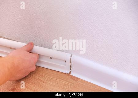 A man installs a floor skirting board. Fixing the plastic skirting board with screws to the wall. Home renovation. Stock Photo