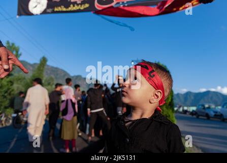 A Kashmiri Shia Muslim boy wearing a headband with Islamic inscriptions looks on towards Shia Muslim men hoisting religious flags and banners as Muharram starts. Muharram is the first month of Islam. It is one of the holiest months on the Islamic calendar. Shia Muslims commemorate Muharram as a month of mourning in remembrance of the Martyrdom of the Islamic Prophet Muhammad's grandson Imam Hussain, who was killed on Ashura (10th day of Muharram) in the battle of Karbala in 680 A.D. Stock Photo