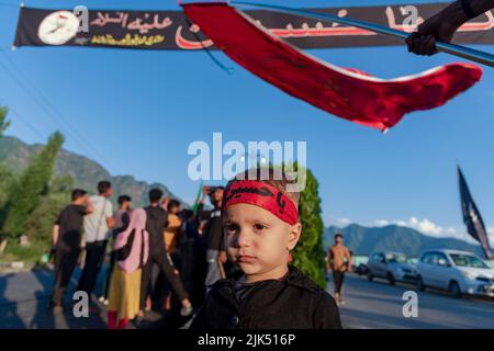 Srinagar, India. 30th July, 2022. A Kashmiri Shia Muslim boy wearing a headband with Islamic inscriptions looks on towards Shia Muslim men hoisting religious flags and banners as Muharram starts. Muharram is the first month of Islam. It is one of the holiest months on the Islamic calendar. Shia Muslims commemorate Muharram as a month of mourning in remembrance of the Martyrdom of the Islamic Prophet Muhammad's grandson Imam Hussain, who was killed on Ashura (10th day of Muharram) in the battle of Karbala in 680 A.D. (Photo by Idrees Abbas/SOPA Images/Sipa USA) Credit: Sipa USA/Alamy Live News Stock Photo