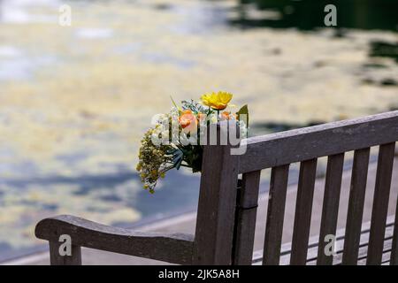 A bouquet of flowers on a park bench next to water. in memory, remembrance. Stock Photo