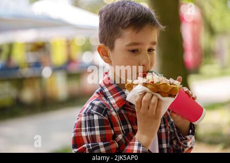 Small Caucasian boy eating sweet waffle at street. Child with fast food in hands. Stock Photo