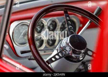 Steering wheel on a custom 1950's Ford F100 pick up truck with stainless steel control panel, dials and red metal coachwork in the background Stock Photo