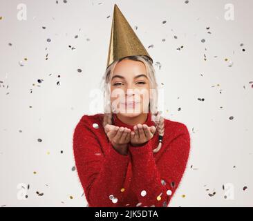 The world has grown weary through the years, but at Christmas it is young. Studio shot of a young woman wearing a party hat and blowing confetti Stock Photo