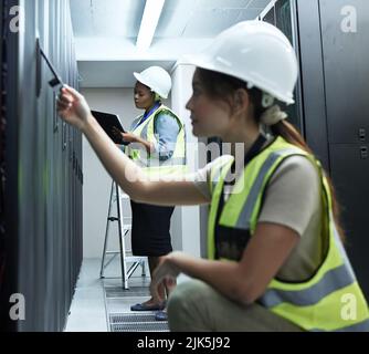 The answer might be behind here. Closeup shot of an attractive young female programmer working in a server room with a colleague in the background. Stock Photo