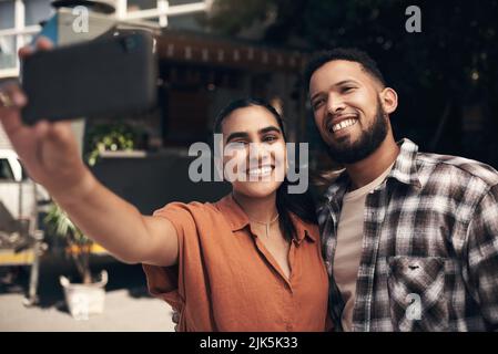 Some much needed bonding time. two young friends standing outside a restaurant and using a cellphone to take a selfie. Stock Photo