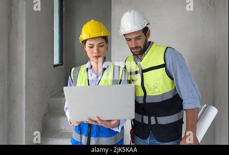 Young engineer with laptop computer explain to foreman about a floor plan. Both wear construction helmet and safety vest. Work environment at the cons Stock Photo