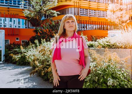 middle aged 40s blonde woman with long hair, in pink scarf and summer clothes, looks at modern architecture with hope and dreaminess. concept of housi Stock Photo