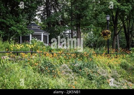 Hillside flower garden with orange daylilies with a gazebo and a lampstand with a hanging petunia flower basket. Stock Photo