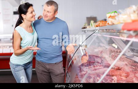 Positive couple customers choosing meat and discussing in butcher’s ...