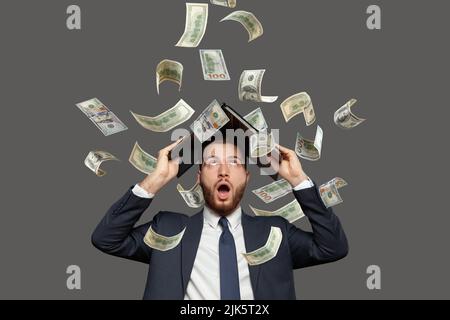 Businessman screaming super excited. Happy young guy celebrating success under money rain falling down dollar bills banknotes isolated gray background Stock Photo