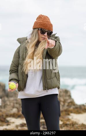 Woman in sunglasses orange beanie and green jacket pointing at camera at Perth dog beach Perth Western Australia Stock Photo