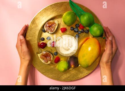Woman's hands serve a beautifully served plate, platter of fresh tropical fruit on a pink table top view. Faceless girl serving on the table. Figs, ma Stock Photo