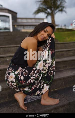 holding her long flower print skirt a brunette latin woman with short black hair, young model sitting on a bleachers, youth beauty and fashion Stock Photo