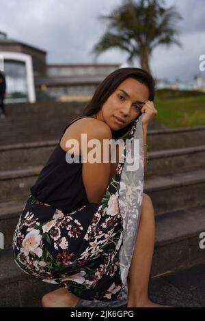 holding up her long flower print skirt a brunette latin woman with short black hair, young model sitting on a bleachers barefoot Stock Photo