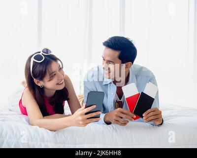 Happy Asian couple with flight booking for summer trip, travel abroad. Young man and woman in casual holding smartphone and two passports and tickets Stock Photo