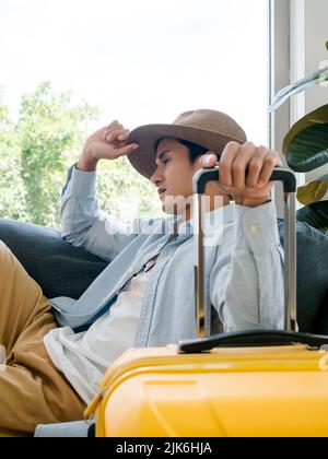 A man with yellow luggage. Handsome Asian male in denim shirt sitting and waiting and thinking while holding suitcase near the glass window, vertical Stock Photo