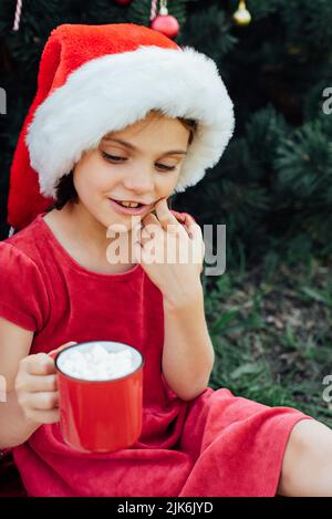 Merry Christmas. Portrait of funny child girl in Santa hat eating gingerbread cookies drinking hot chocolate outside having fun. Happy Holidays. kid e Stock Photo