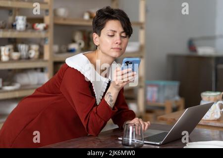 Young female ceramic artisan holding mobile phone using social media to communicate with customers Stock Photo
