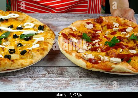 Chef puts on table two homemade pizza with cheese, sausage, chicken meat, olives and basil leaves. Tasty unhealthy fast food snack of Mediterranean fo Stock Photo