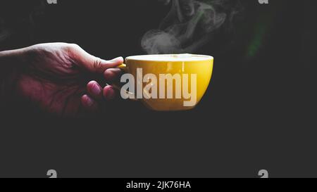 Close-up isolated hand holding yellow mug on black background. Hand hold simple yellow cup. Perfect for both tea and coffee. Stock Photo