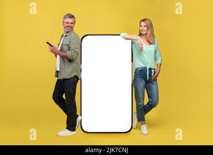 Great app. Full body length of middle aged couple leaning on big smartphone with white empty screen Stock Photo